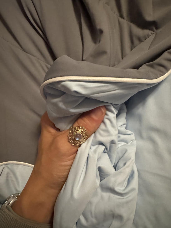 A hand holding the piped edge of a comforter to show its dark gray outer fabric and pale blue reverse.