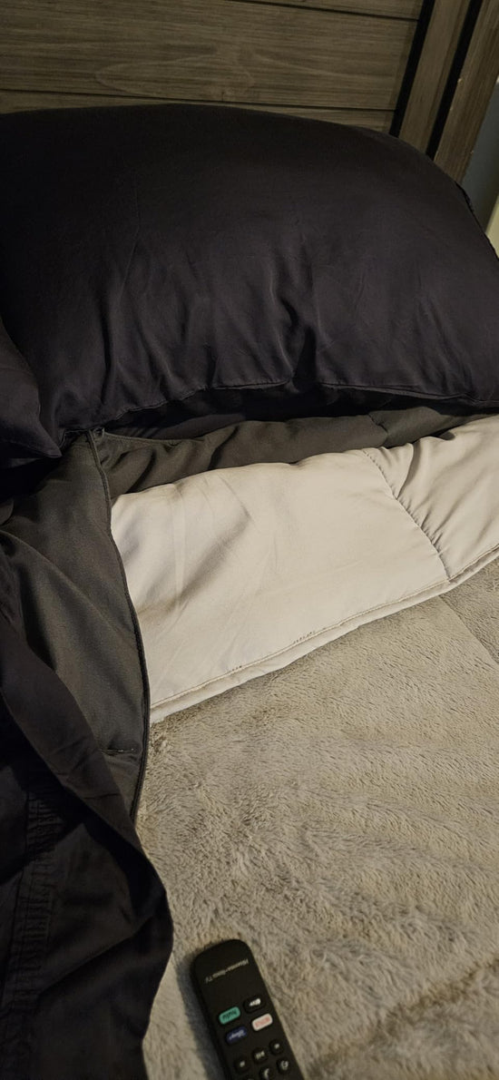 Dark pillow and sheet partly pulled back over a light mattress with a remote control near the foot.