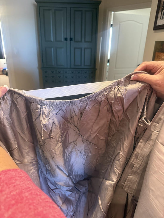 Hands holding up the elastic edge of a wrinkled mauve fitted sheet in a bedroom.