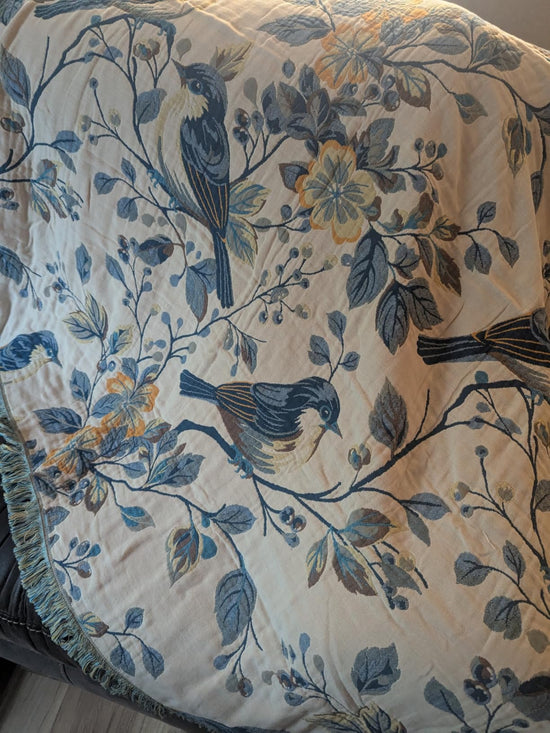 Cream throw covered with blue birds and flowering branches, with a fringed edge visible below.