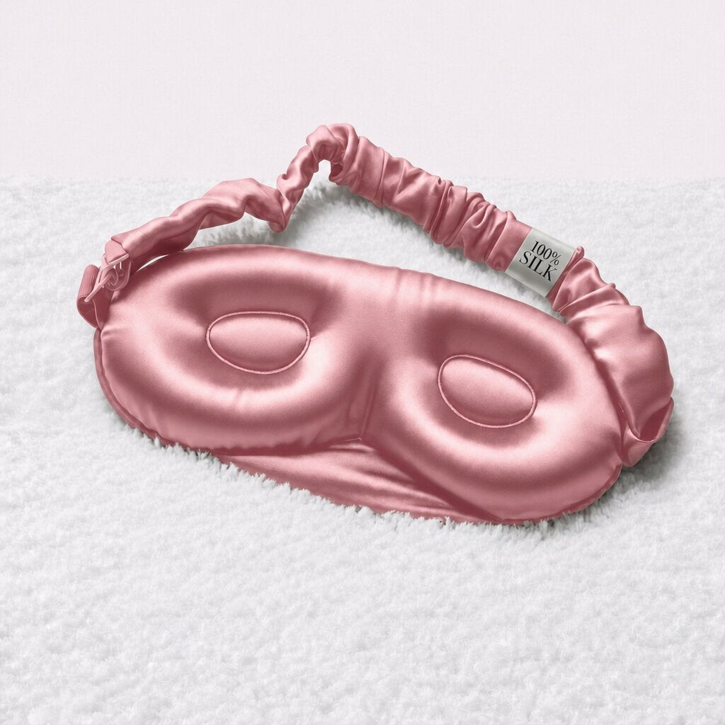 Inside of a rose pink contoured sleep mask resting on a white towel, showing recessed eye cups and a gathered adjustable strap.