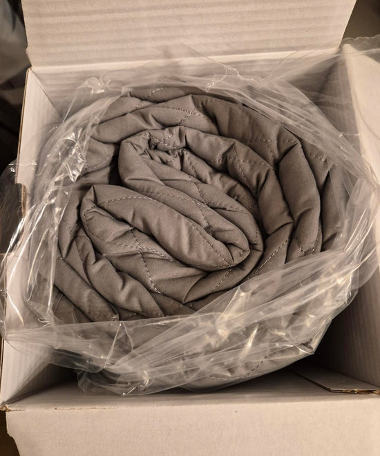 Rolled gray quilt inside a clear plastic bag in an open cardboard box.