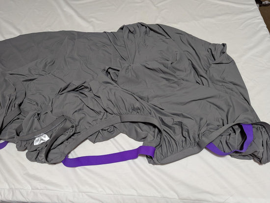 Gray fitted sheet spread out on a bed with purple corner straps and a white fabric label visible.