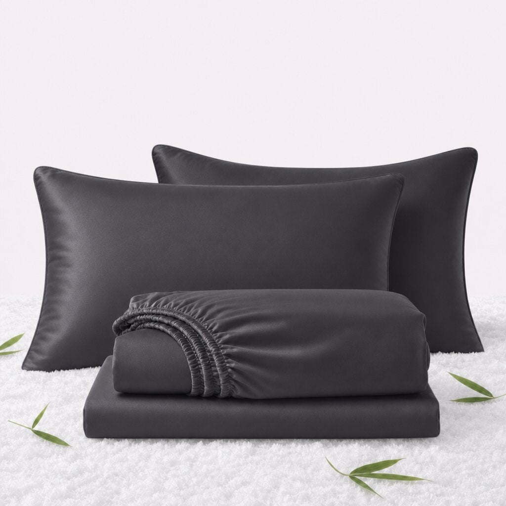 Folded charcoal gray sheets in front of 2 matching pillows on a white textured surface with scattered bamboo leaves.