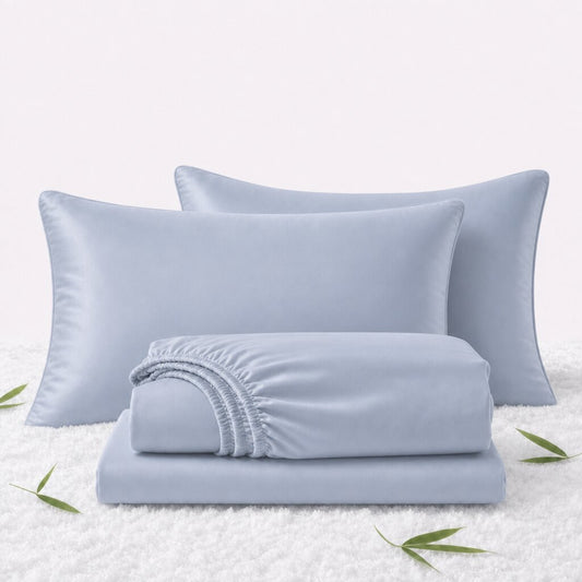 Folded light blue sheets in front of 2 matching pillows on a white textured surface with scattered bamboo leaves.
