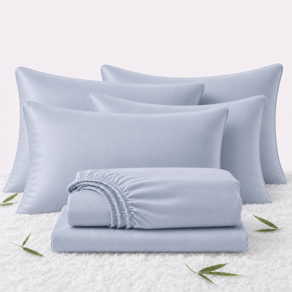 Folded light blue sheets in front of 4 matching pillows on a white textured surface with scattered bamboo leaves.