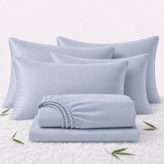 Folded light blue sheets in front of 4 matching pillows on a white textured surface with scattered bamboo leaves.