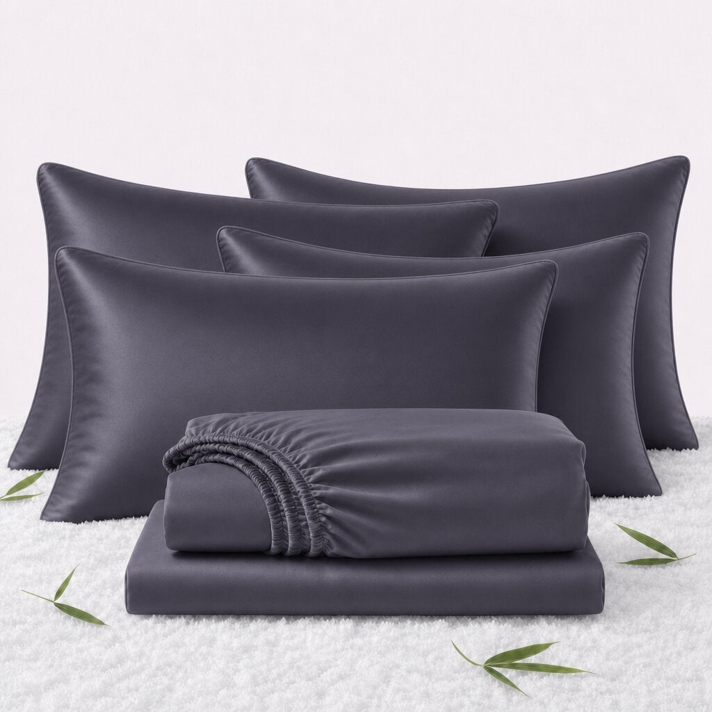 Folded dark gray sheets in front of 4 matching pillows on a white textured surface with scattered bamboo leaves.