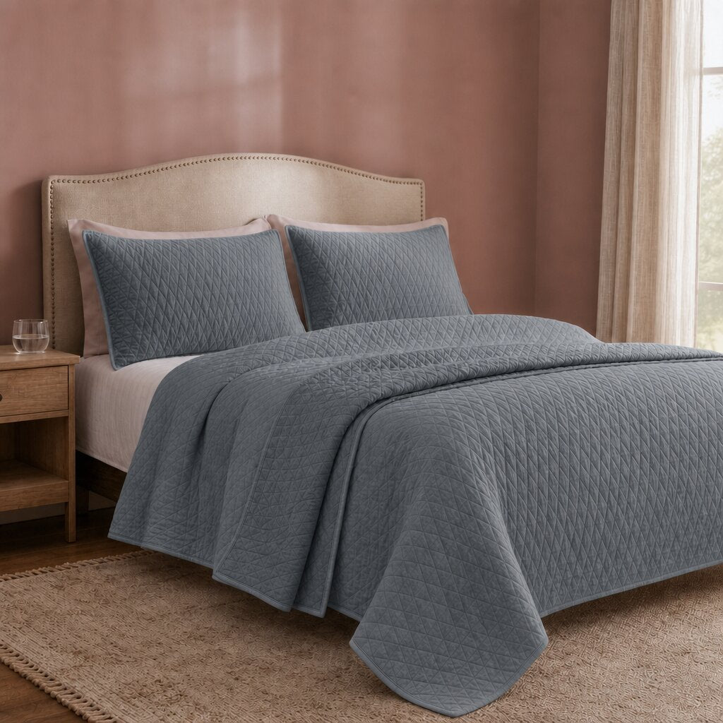 Blue gray diamond quilt and two matching shams on a cream upholstered bed in a softly lit bedroom.