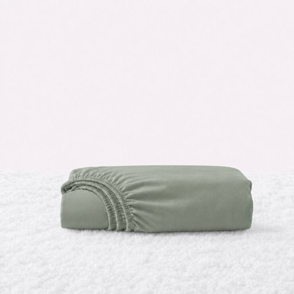 Folded sage green fitted sheet with its gathered elastic edge visible on a white textured surface.