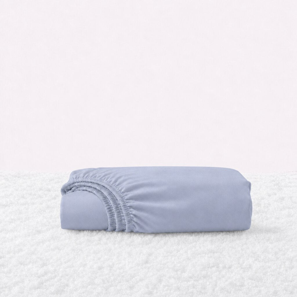 Folded pale blue gray fitted sheet with its gathered elastic edge visible on a white textured surface.
