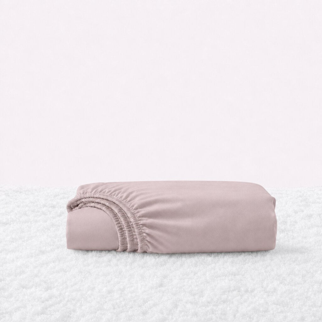 Folded blush pink fitted sheet with its gathered elastic edge visible on a white textured surface.