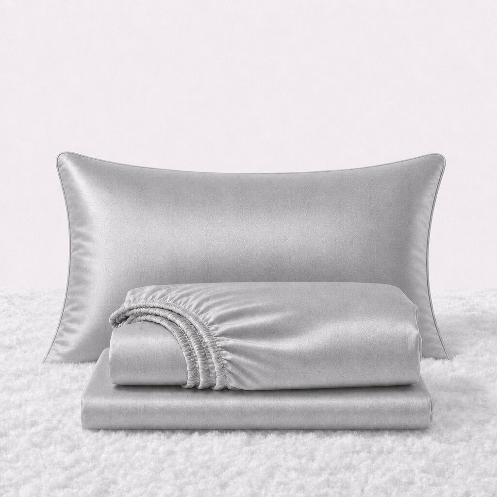 Folded shiny silver gray satin sheets in front of one matching pillow on a white textured surface.