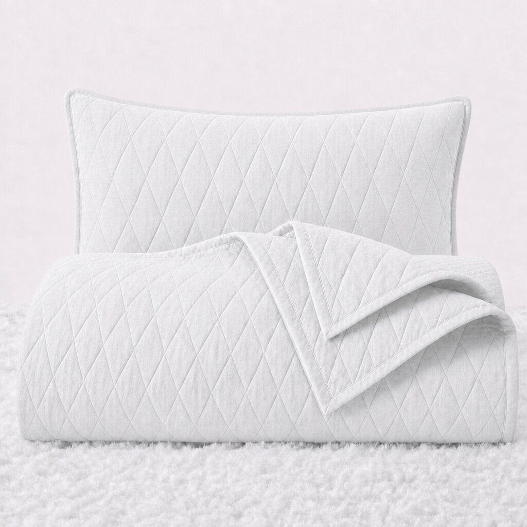 Folded white diamond stitched quilt with one matching pillow sham standing behind it on a white textured surface.