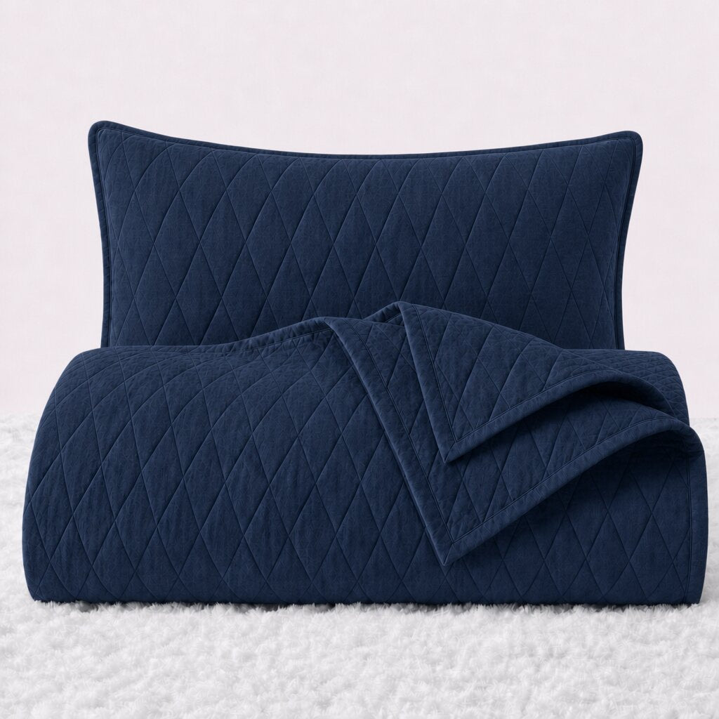 Folded navy blue diamond stitched quilt with one matching pillow sham standing behind it on a white textured surface.