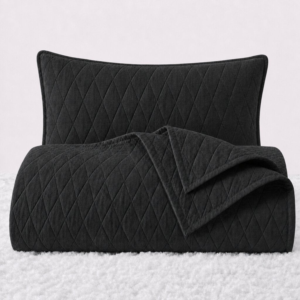 Folded black diamond stitched quilt with one matching pillow sham standing behind it on a white textured surface.
