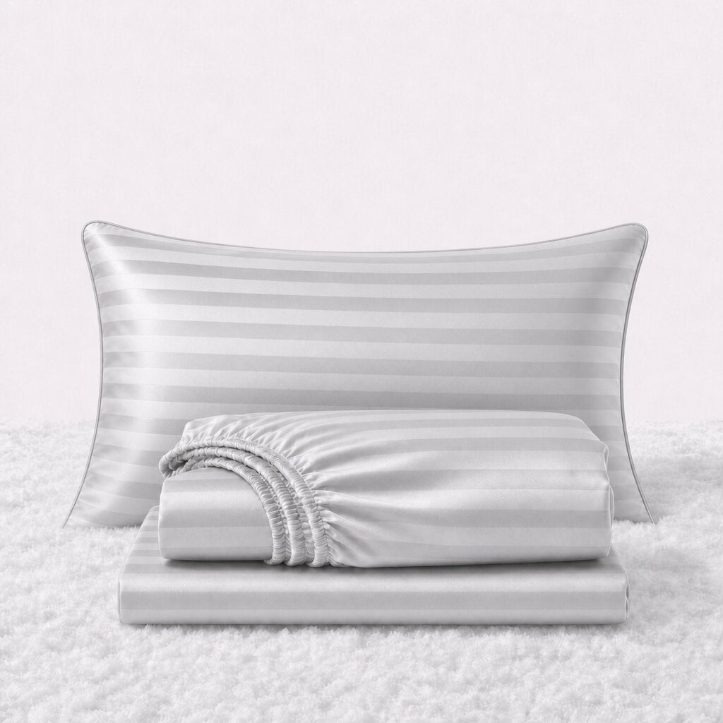 Folded shiny silver gray striped satin sheets in front of one matching pillow on a white textured surface.