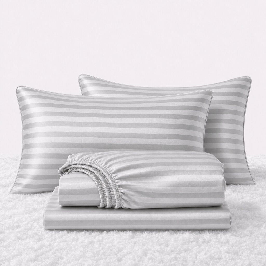 Folded shiny silver gray striped satin sheets in front of two matching pillows on a white textured surface.