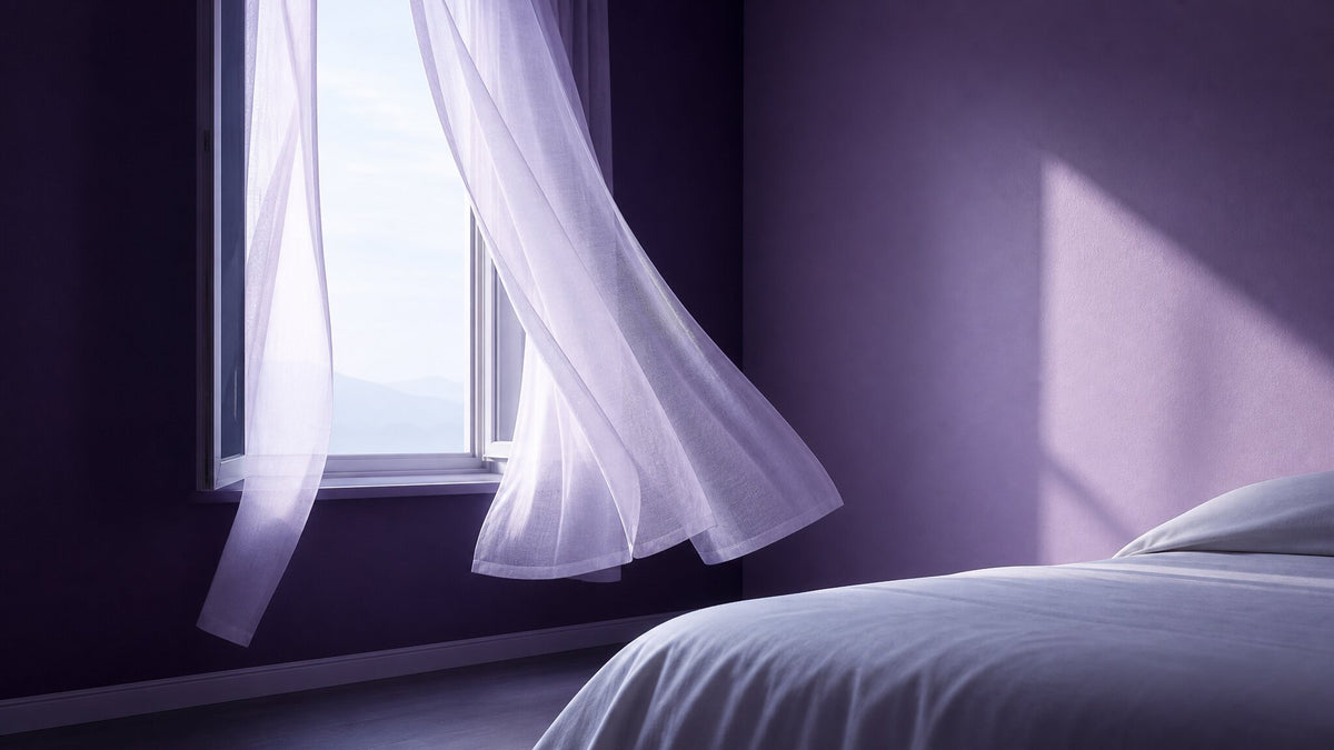 Sheer white curtain billowing through an open window beside a bed in a sunlit purple bedroom.